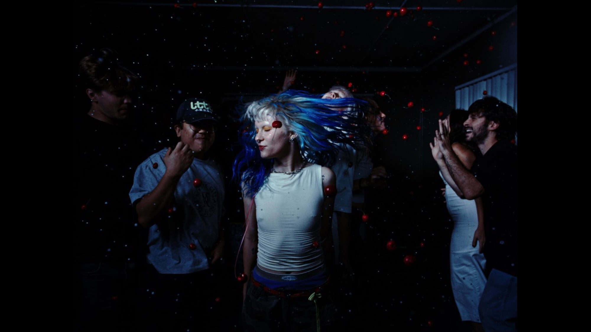 A group of people dance in a dark room; at the center, a person with blue-tinted hair moves in rhythm, while small red balls float in the air—a scene reminiscent of spring festivals or film festivals in Seattle.