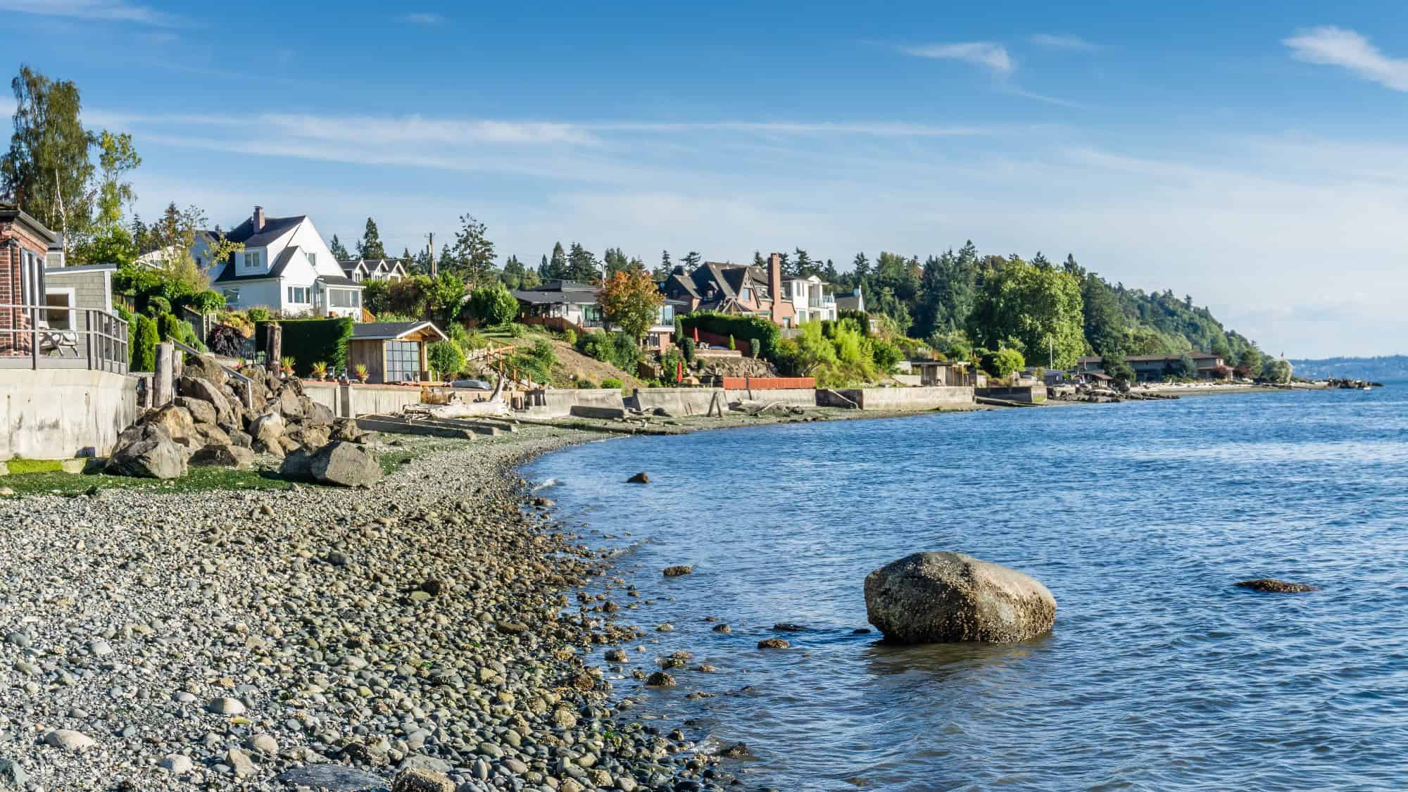Houses line a rocky shoreline beside calm blue water under a clear sky, with trees and greenery in the background.