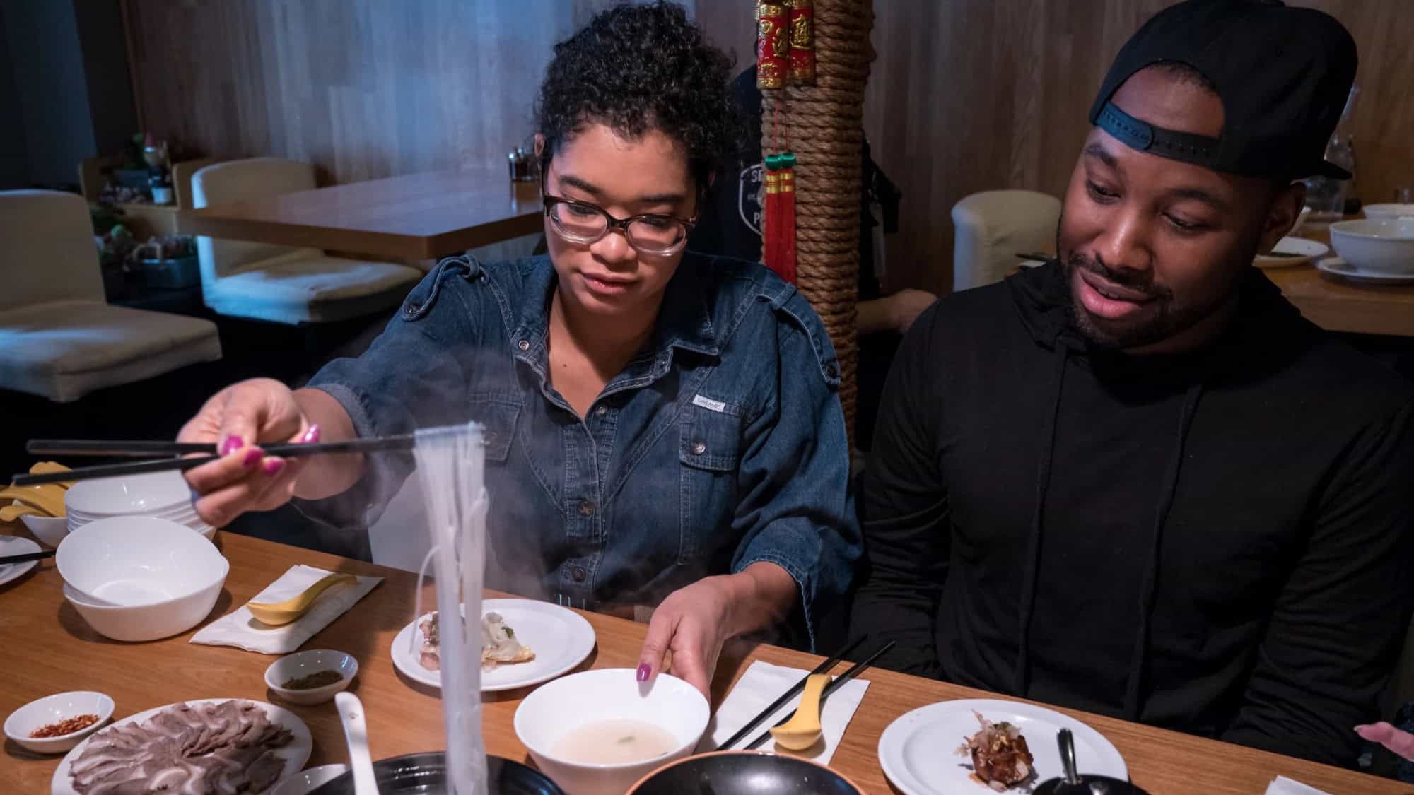 Two people sit at a restaurant table with various dishes; one person uses tongs to serve noodles into a bowl while the other looks on.