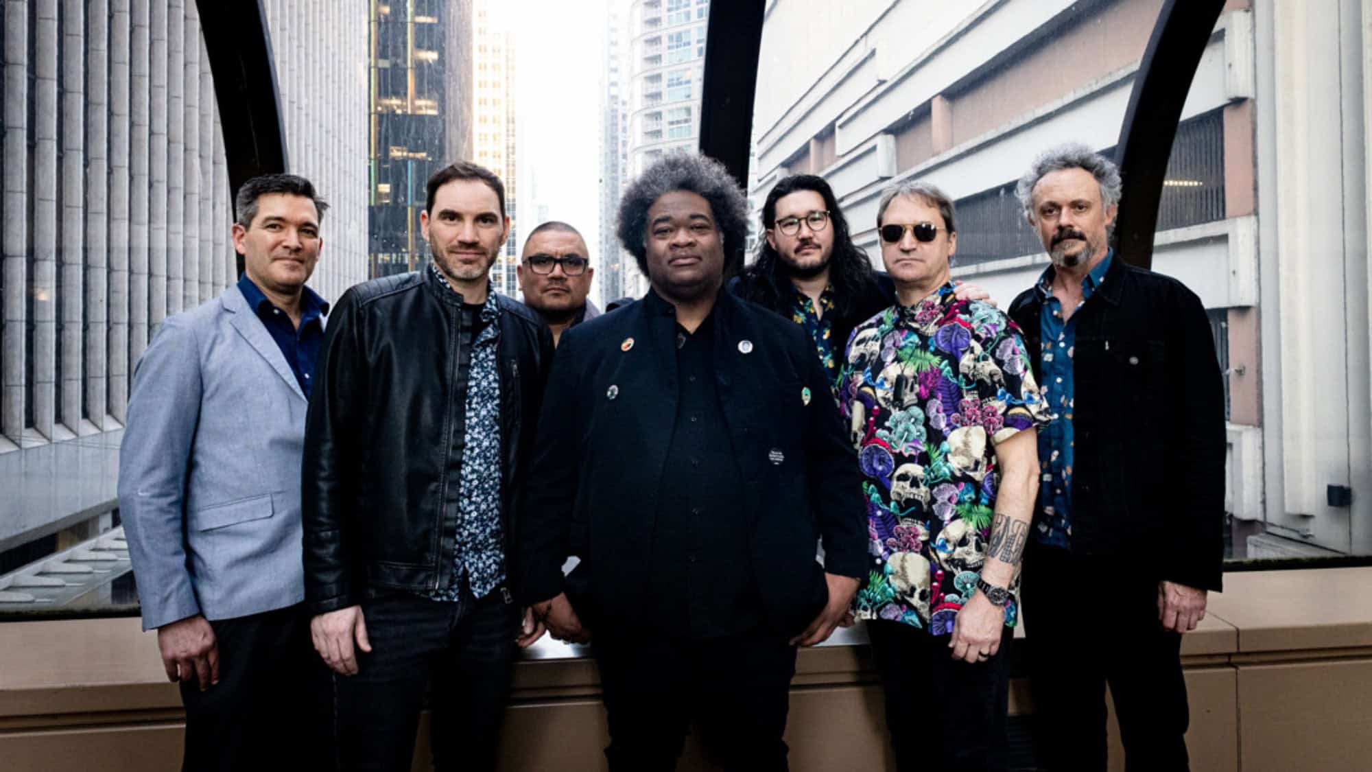 Seven men stand side by side in front of city buildings, wearing casual and patterned clothing, posing for a group photo within a covered walkway.