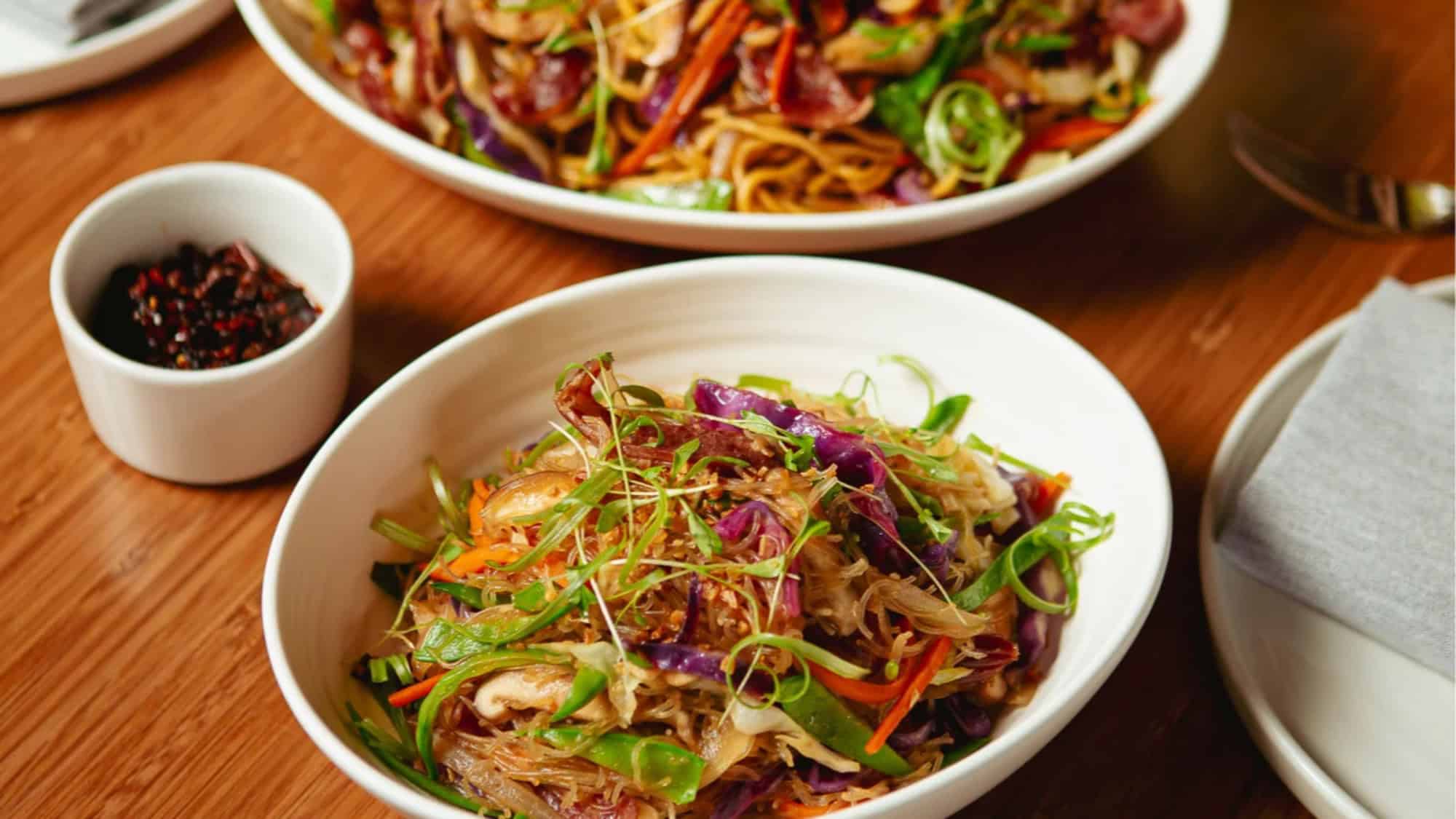 Two bowls of stir-fried noodles with vegetables and greens on a wooden table, accompanied by a small dish of sliced red chili peppers.