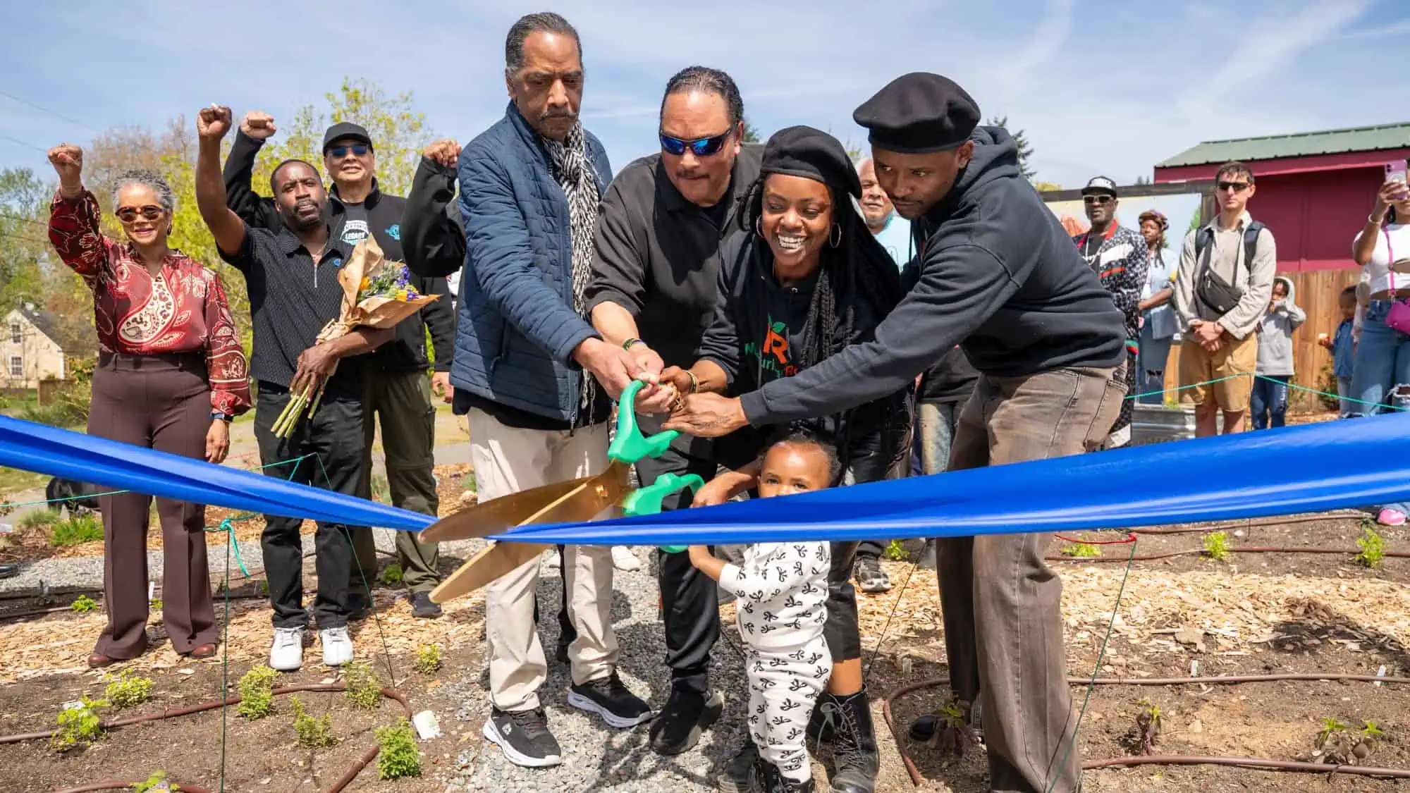 A group of people, including a child, cut a blue ribbon with large scissors at an outdoor event, while others watch and cheer in the background.