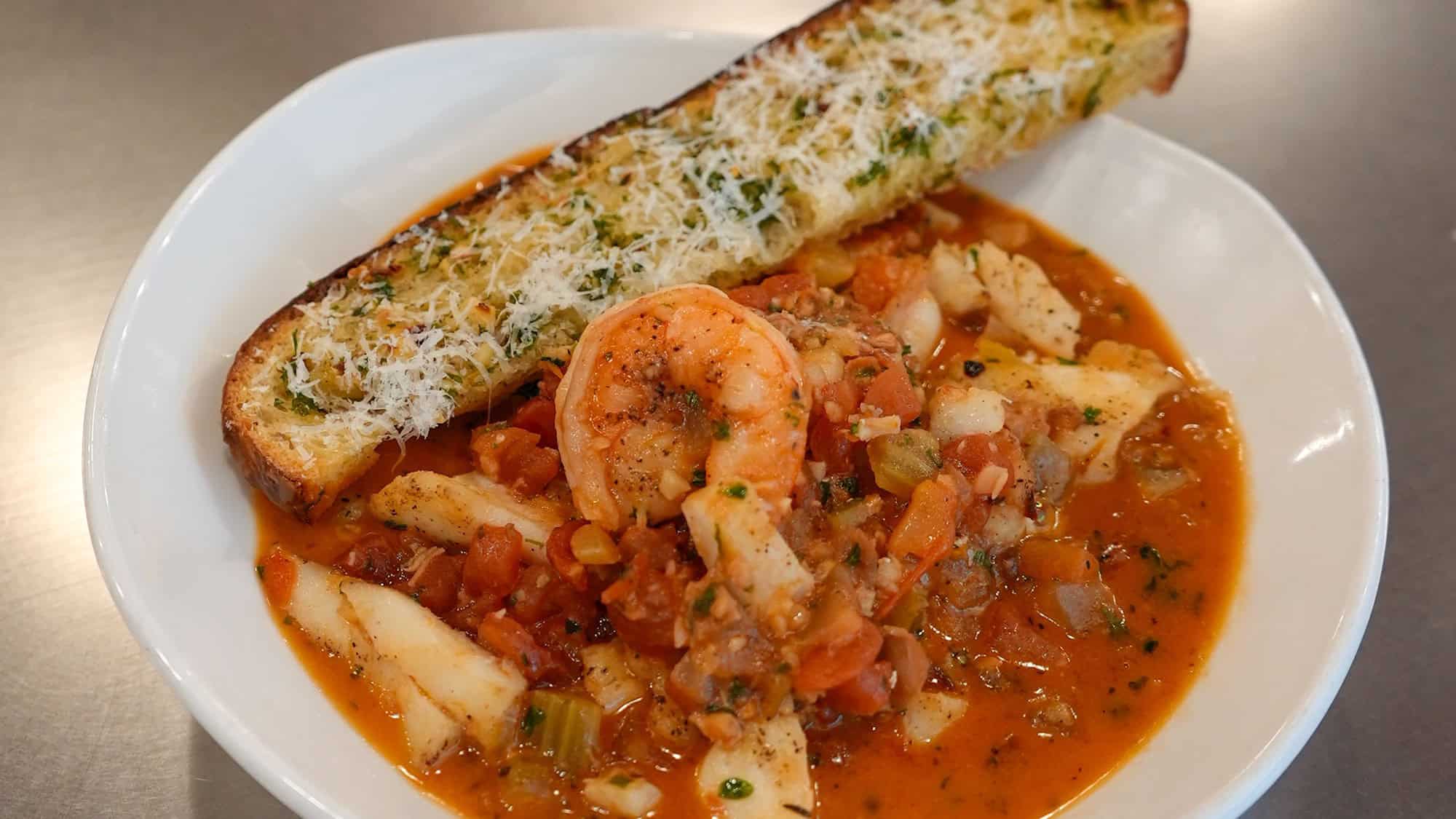 A bowl of seafood stew with shrimp and fish in tomato broth, served with a slice of garlic bread topped with grated cheese—perfect for spring food lovers seeking fresh April recipes.