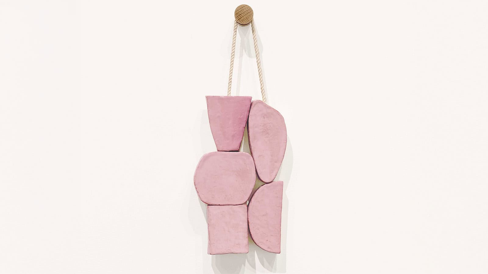A wall-mounted sculpture by Gabriel Stromberg made of six irregular pink clay pieces, stacked and suspended by a rope looped over a wooden peg against a white background—a highlight from recent Studio Sessions.