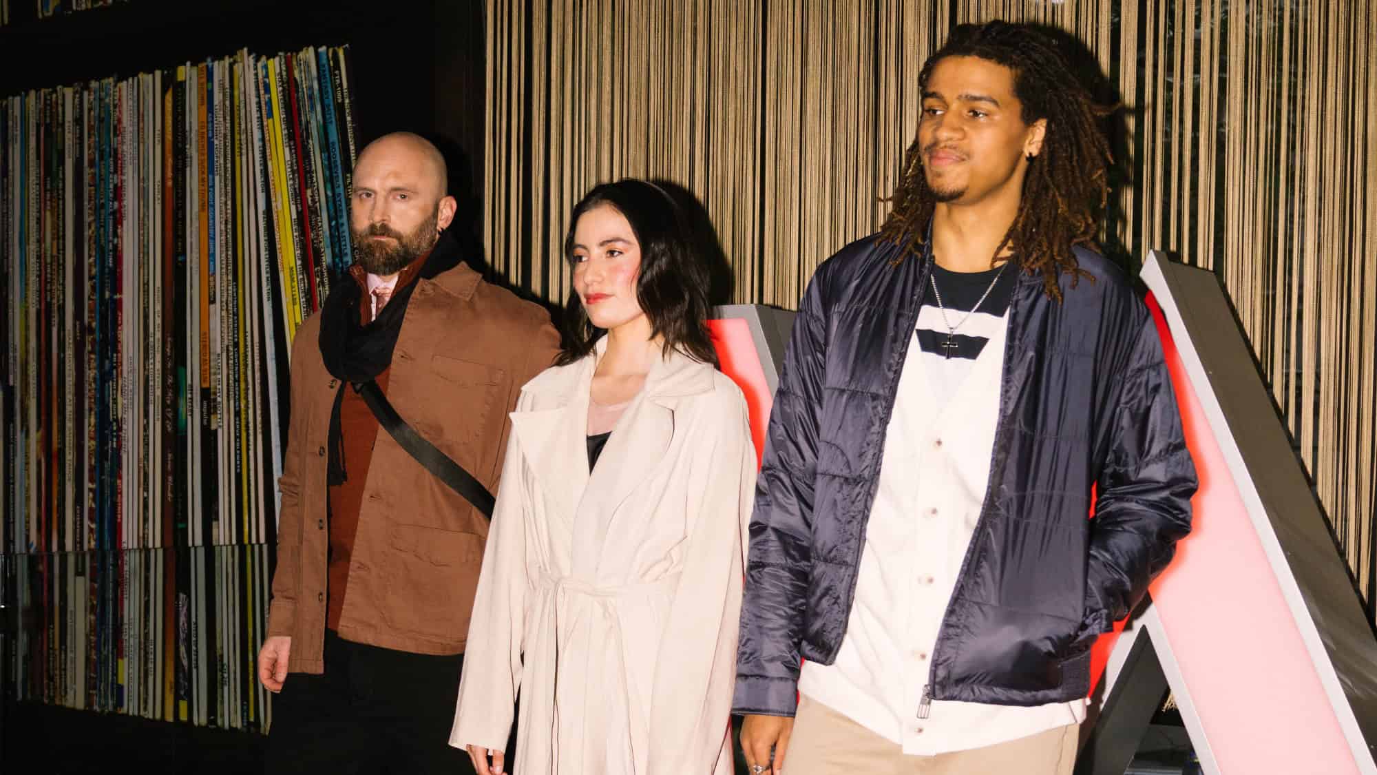 Three people stand side by side indoors, in front of shelves filled with books or records and vertical blinds. Dressed as if for a fashion show, they wear casual jackets and have neutral expressions.