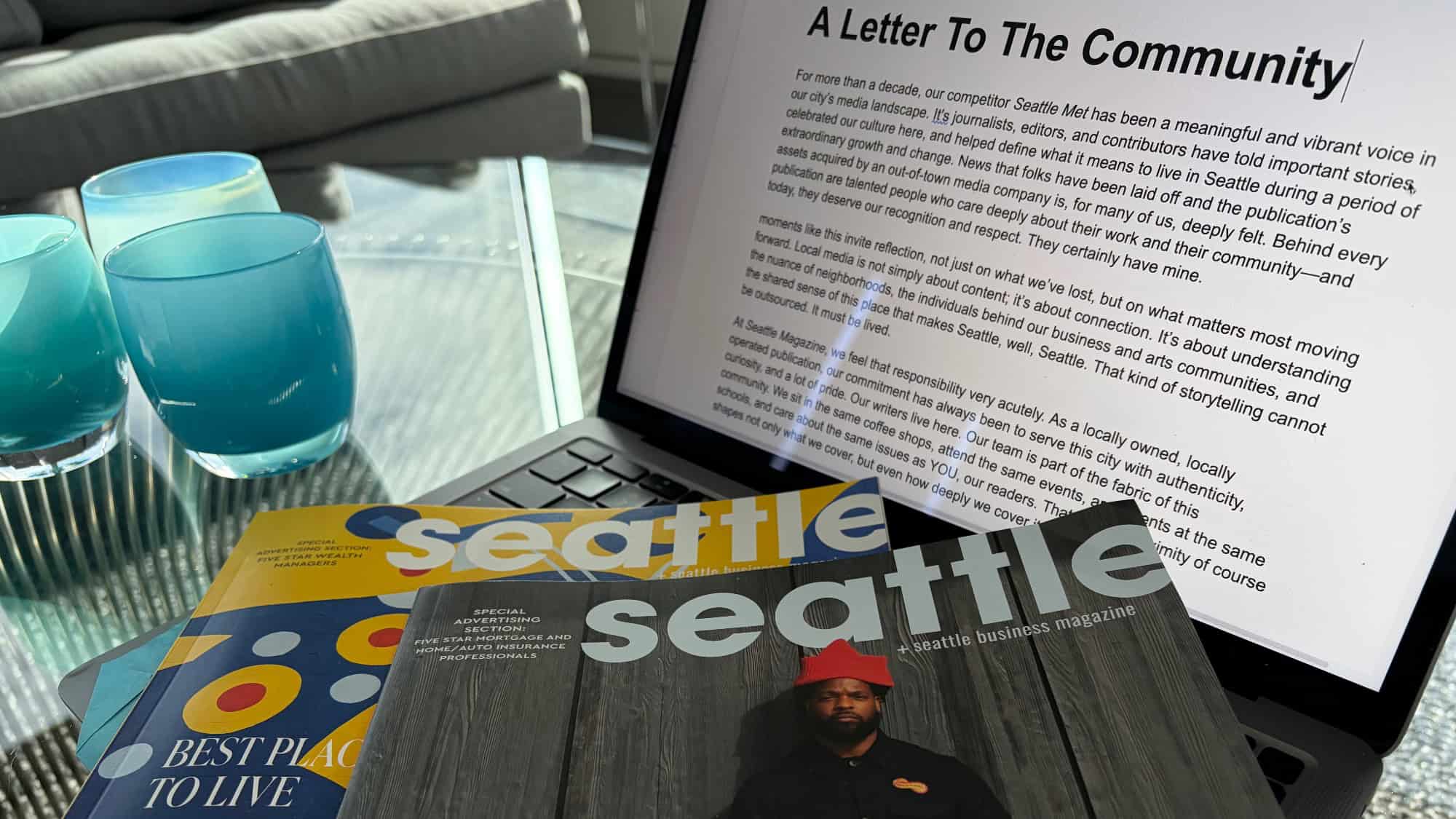A laptop displays a letter to the community, alongside two Seattle magazine issues and a glass table with three blue glasses in the background.