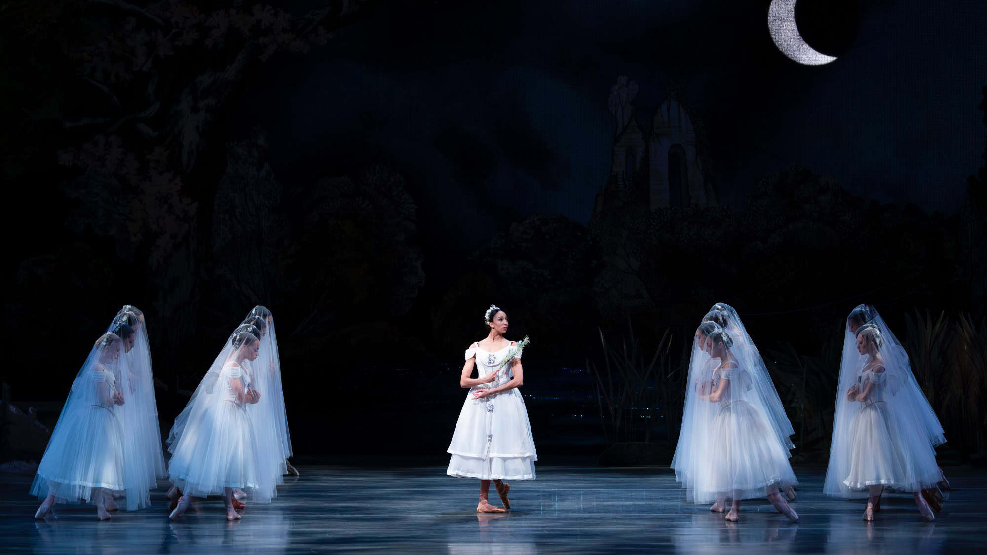 A ballerina in a white costume stands center stage, surrounded by dancers in white dresses and veils, beneath a crescent moon in a dark, forested setting that evokes the elegance of ballet performed at a sculpture park.