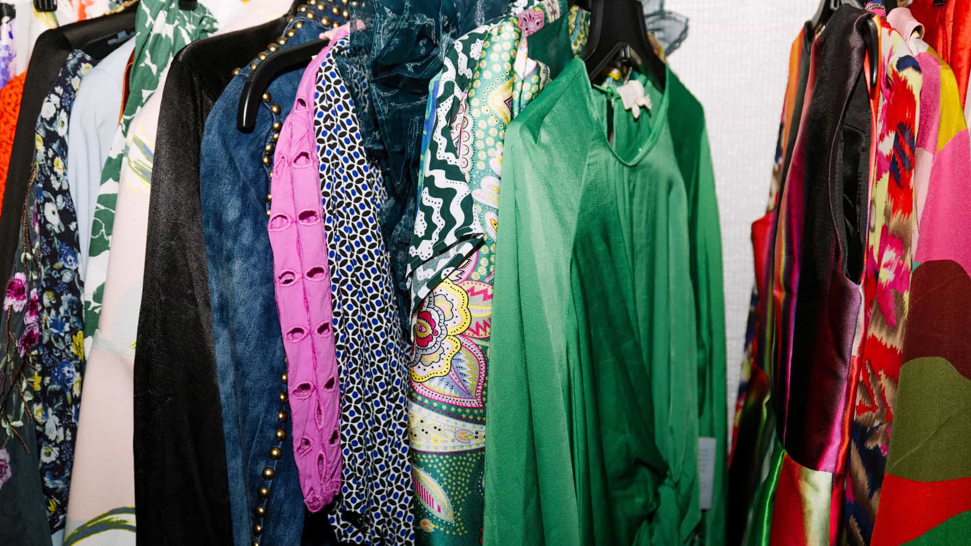 A rack of colorful, assorted clothing including dresses, shirts, and jackets in various patterns and fabrics hanging on black hangers.