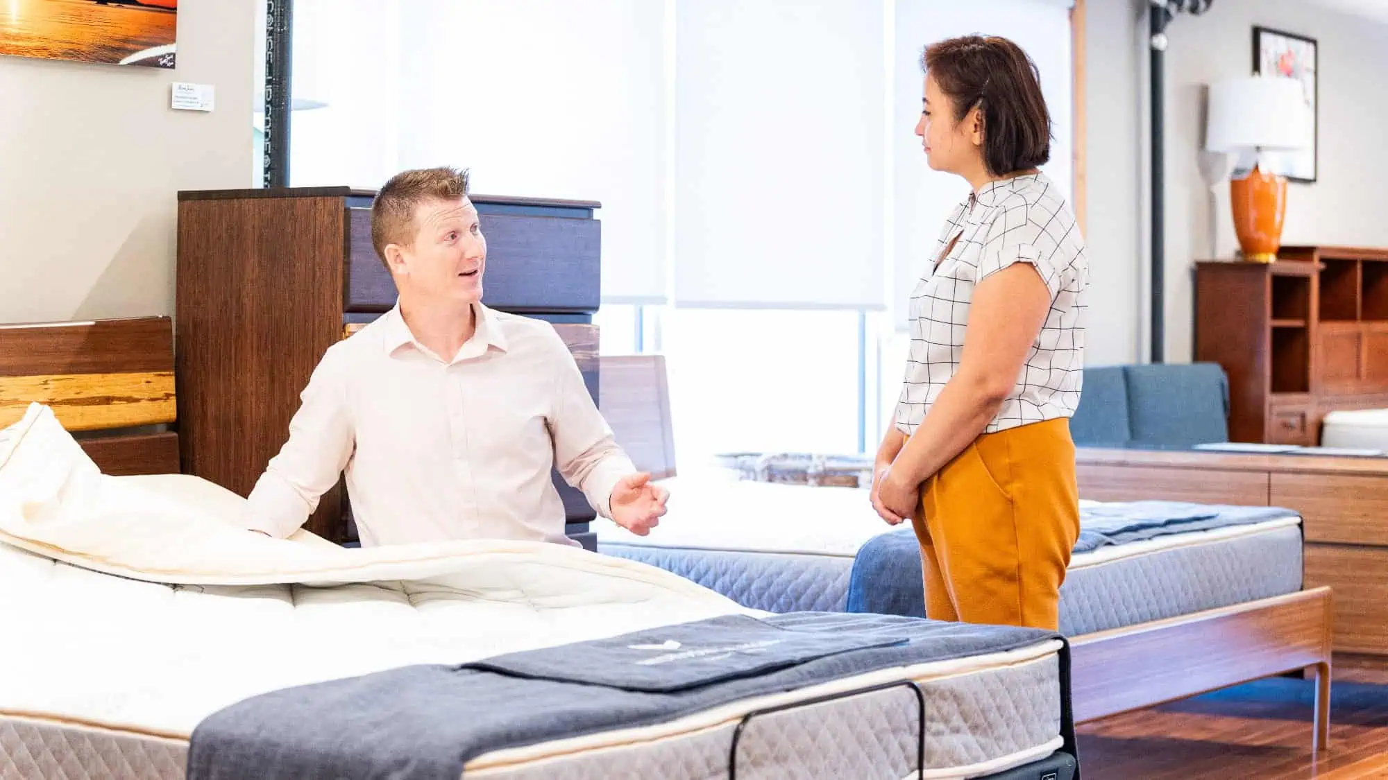 A man sitting on a mattress talks to a woman standing beside him in a brightly lit, unique mattress store.