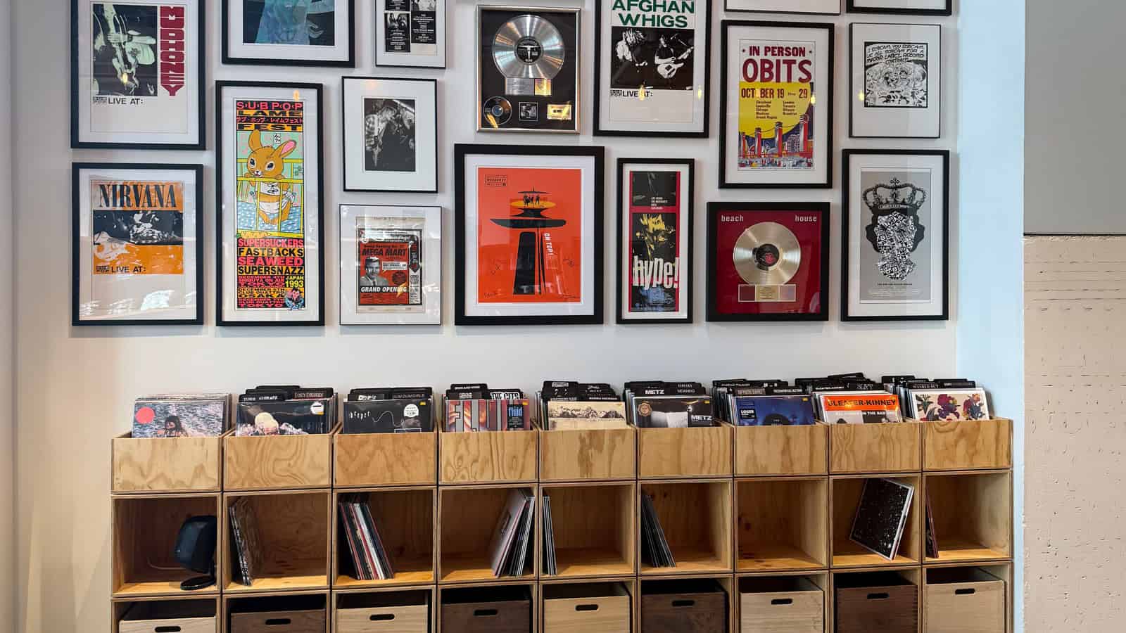 Wall with framed music memorabilia and posters displayed above wooden shelves holding vinyl records in a store or music-themed space.