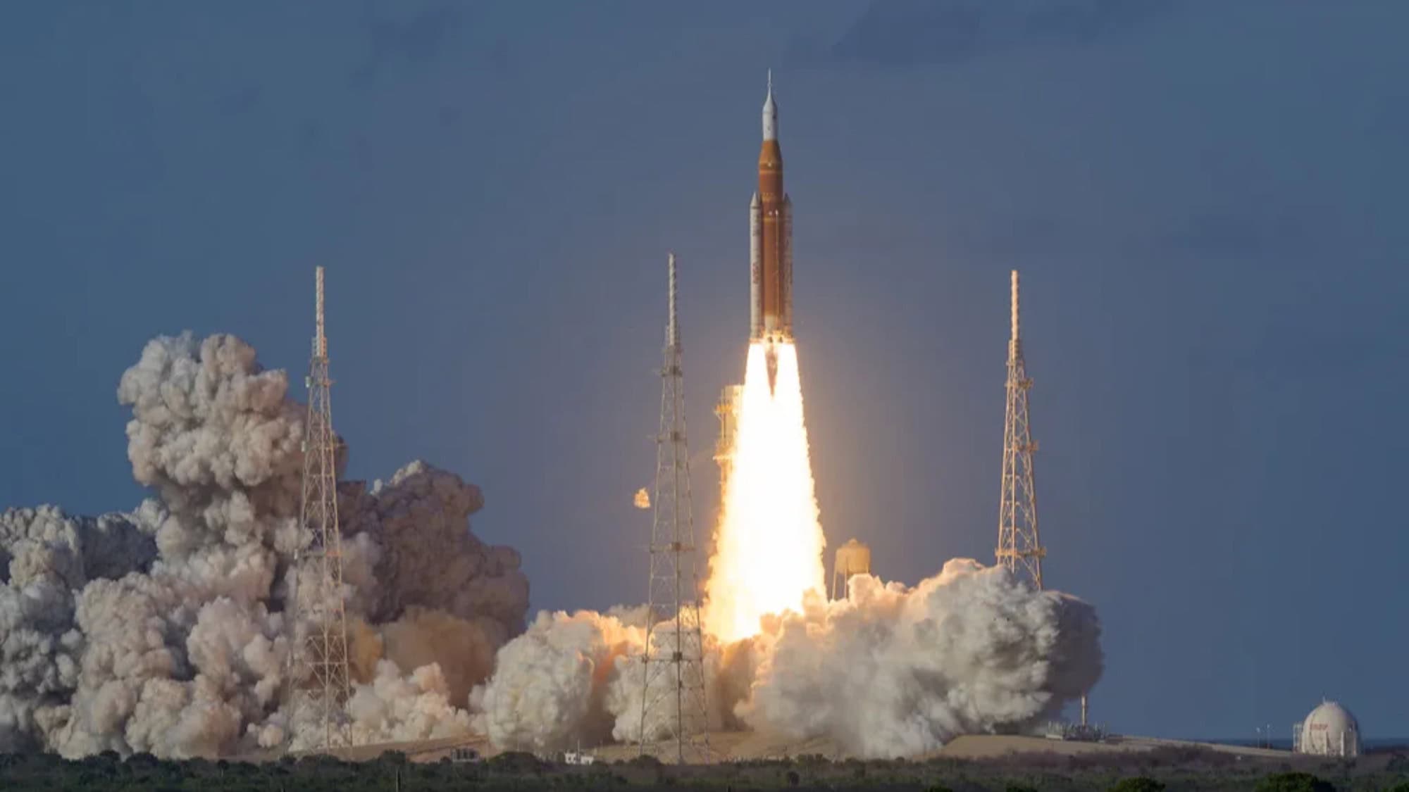 A rocket launches into the sky from a launch pad, surrounded by towers and clouds of smoke against a blue sky.