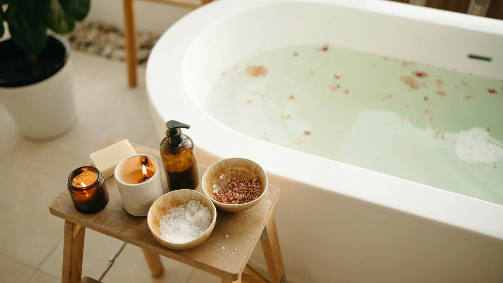A white bathtub filled with water and floating petals is next to a wooden stool holding a lit candle, a soap bar, a pump bottle, and bowls with bath salts and dried flowers.