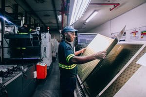 Printer Thabo Mnisi inspects a sheet of 729 printed metal crowns, prior to varnishing, pressing out, and the final stage of lining.