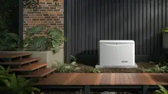 A Kohler 26kW Generator outside a home with dark vertical siding near a wooden walkway and wooden steps with plants on both sides