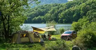 A rustic campsite on the shore of a mountain lake. A yellow tent with sleeping area and front room. A dining fly covers the entrance while a couple sits at a table on camp chairs.Their car is nearby, and a bicycle with a basket in front stands ready for a ride. The trees around the lake are green, and the mountain foliage in the background is blue-green.