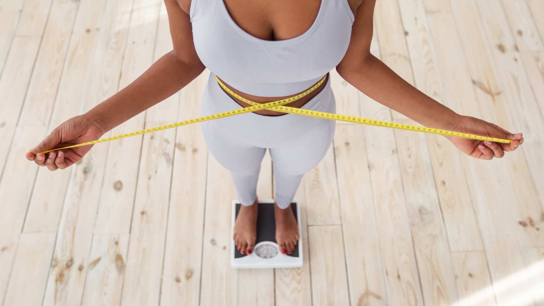 above-view-of-african-american-lady-measuring-her-waist-with-tape-standing-on-scales-indoors-closeup-young-black-woman-showing-results-of-slimming-diet-or-liposuction-promoting-healthy-living