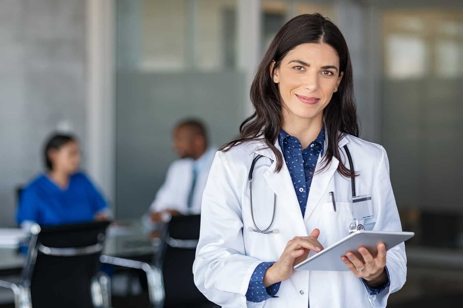 portrait-of-beautiful-mature-woman-doctor-holding-digital-tablet-and-looking-at-camera-confident-female-doctor-using-digital-tablet-with-colleague-talking-in-background-at-hospital-latin-nurse-in-la