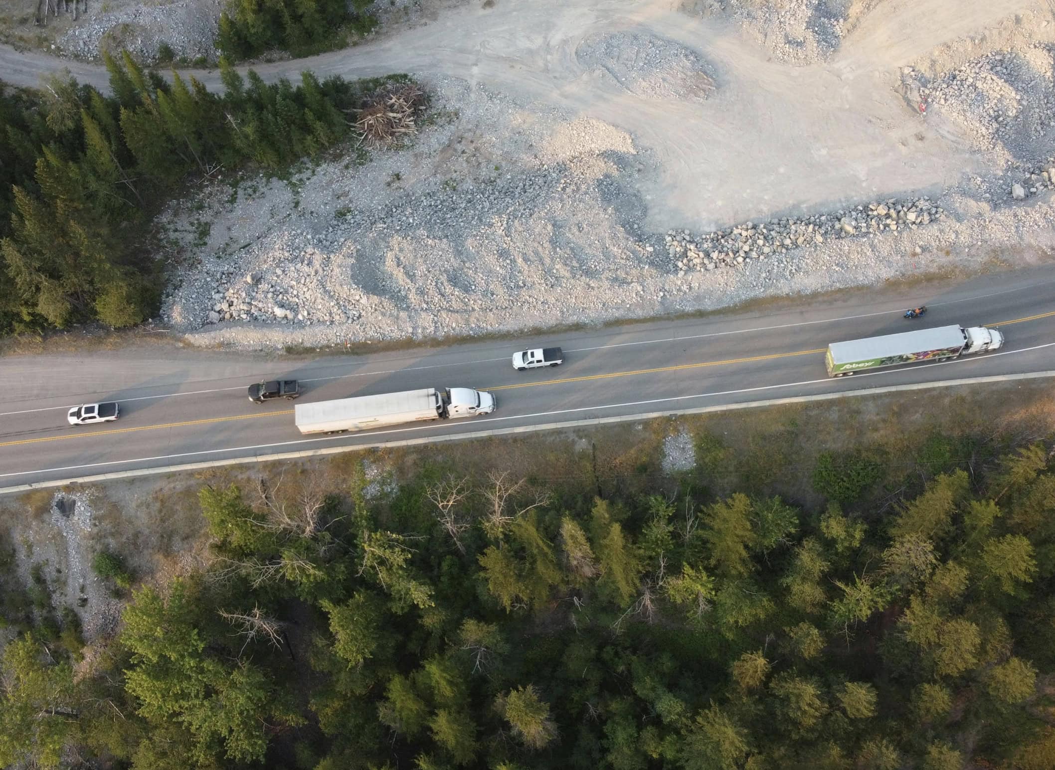 Aerial view of a two-lane highway with semi-trucks and cars traveling between a rocky excavation site and a dense forest.