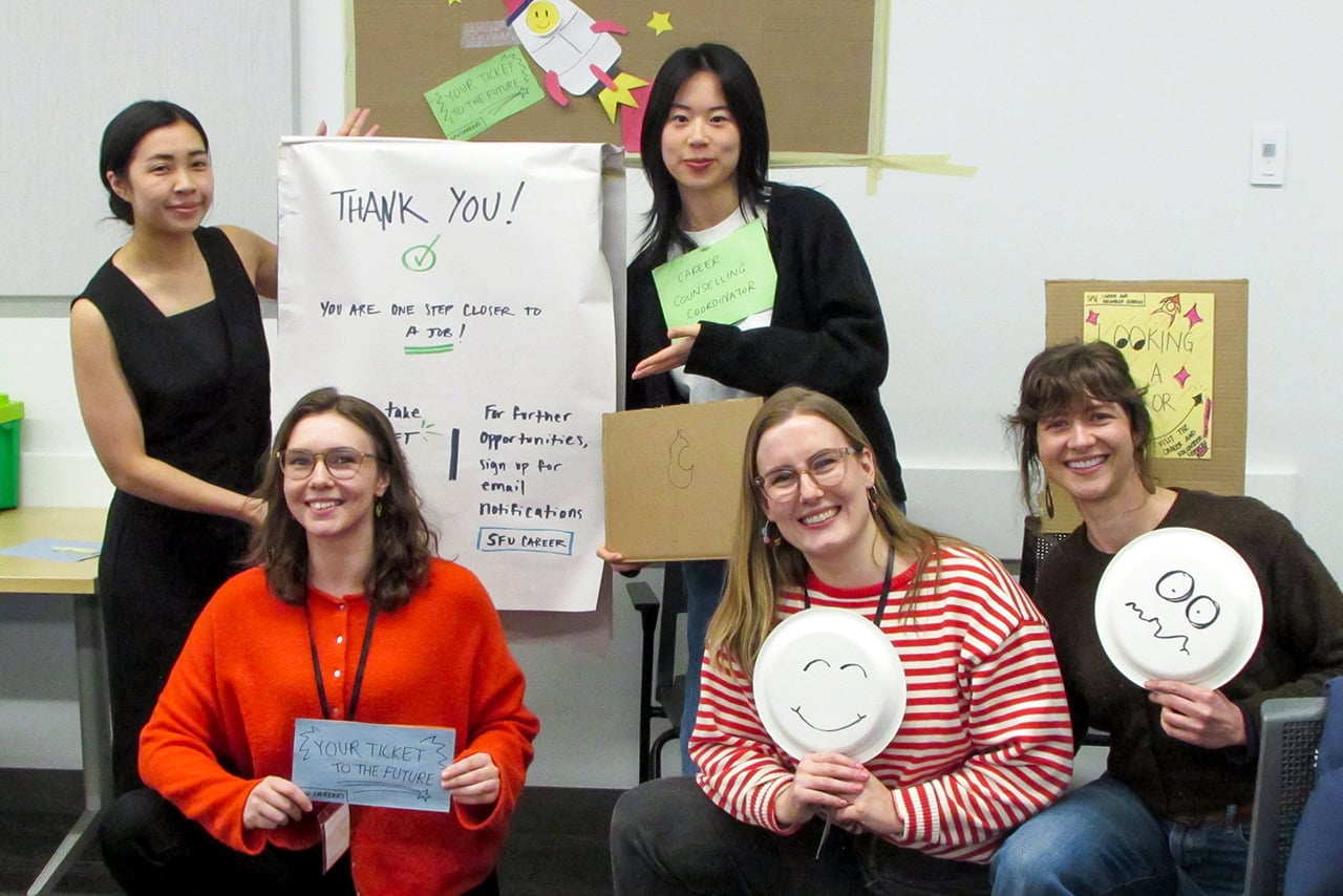 Five smiling participants posing with handmade signs, cardboard props, and paper plates with faces drawn on demonstrating different emotions.