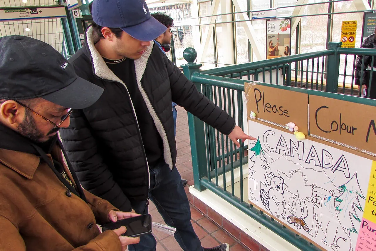 Two participants at a skytrain station pointing at a hand-drawn poster with a heading of "Please Colour Me" taped to a railing.