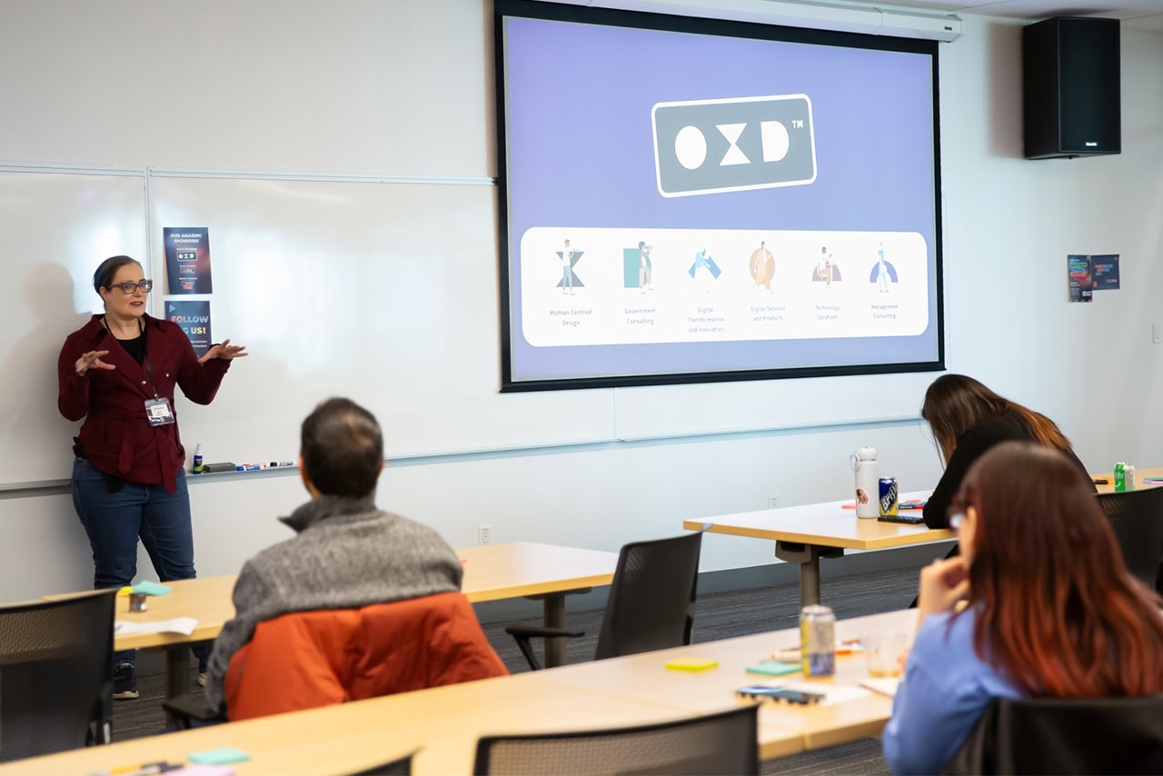 OXD's Deb MacKenzie presenting to an attentive audience at the Vancouver Service Jam 2026, with a large screen displaying the OXD logo and OXD's services icons beside her.