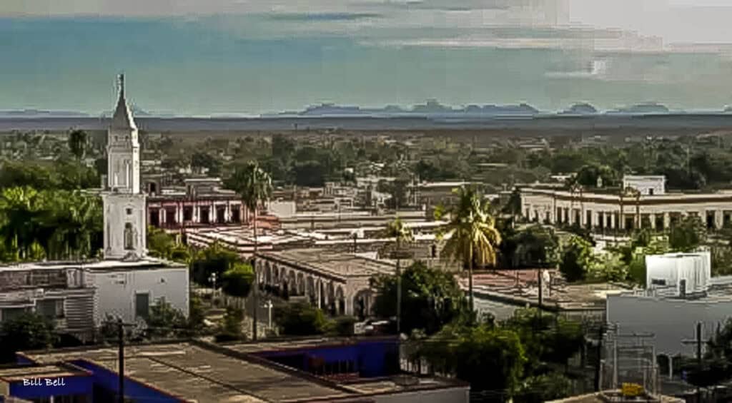 "El Fuerte, Sinaloa"The charming colonial town of El Fuerte, Sinaloa, viewed from above, showcases its historic architecture and lush surroundings. The town's prominent white church tower stands tall against the backdrop of the distant Sierra Madre Occidental mountains, highlighting El Fuerte's blend of rich history and natural beauty.
