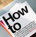 A collection of design books on a couch, showcasing 'How to' by Michael Bierut